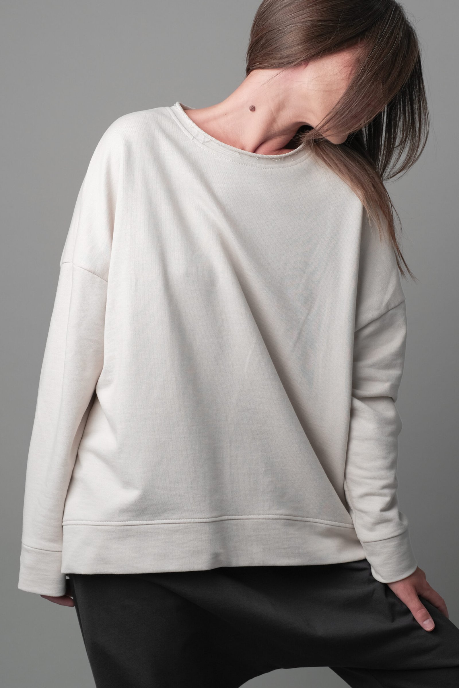 Unisex sweatshirt showcased in a neutral pose with a focus on the relaxed and oversized fit, featuring dropped shoulders. The garment has a loose-fitting raw-edge neckline, straight hem and cuffs, with a slightly longer hem at the back. The model stands against a plain grey background that highlights the sweatshirt's design and comfortable silhouette.